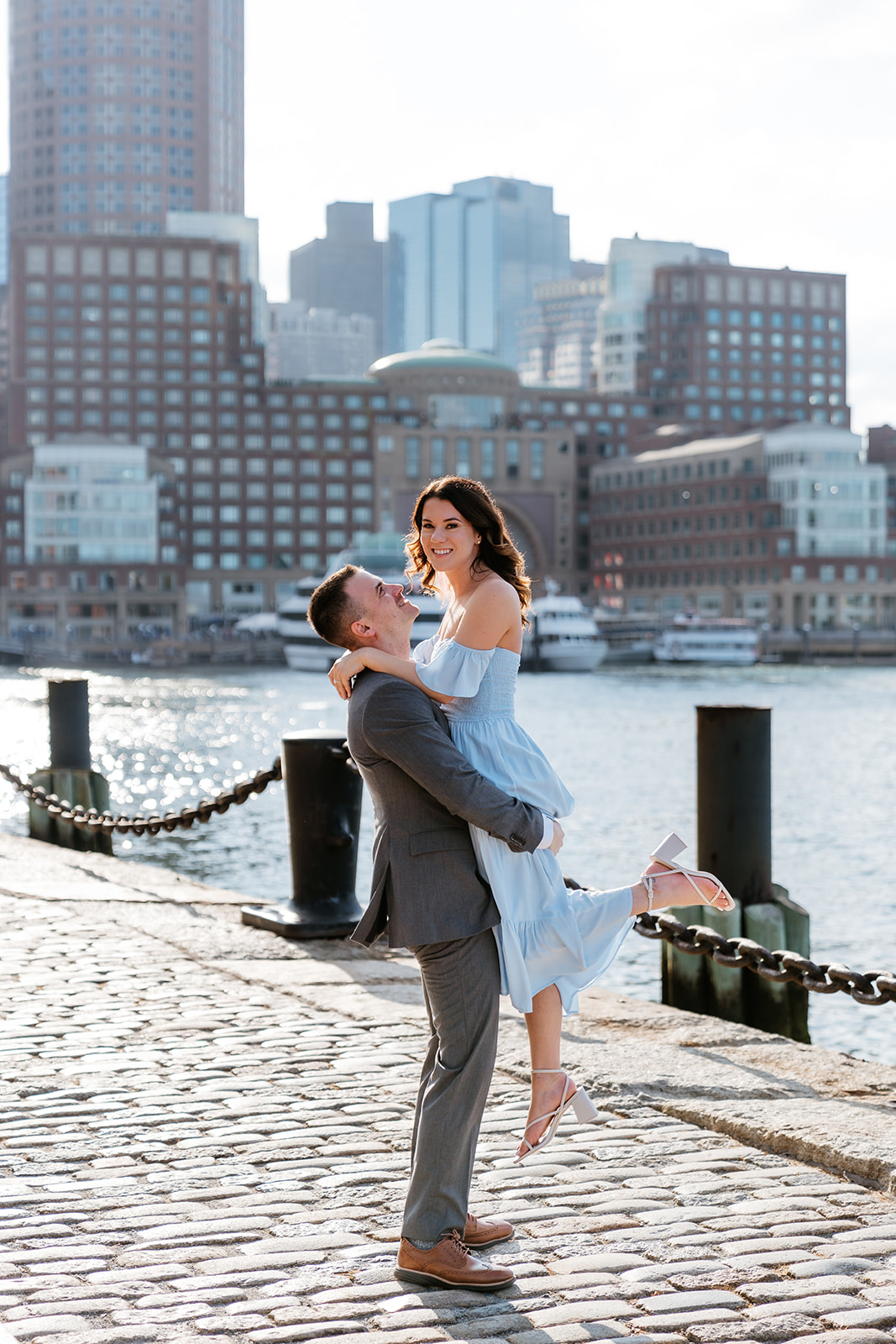 Couple embracing on a city waterfront with skyscrapers in the background at Fan Pier Park as they take Boston engagement photos