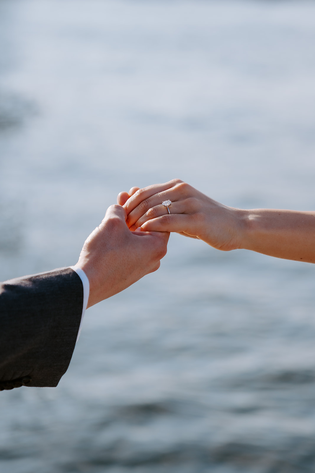 Two people holding hands, one wearing an engagement ring against a water backdrop.
