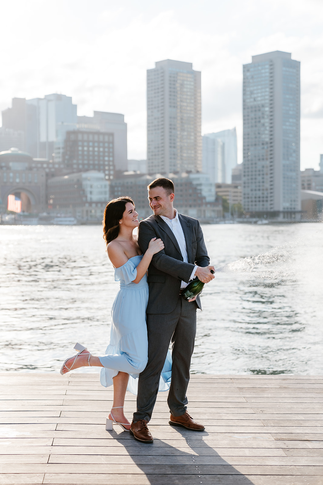 A couple embracing on a wooden pier with a city skyline in the background at Fan pier park as they take Boston engagement photos