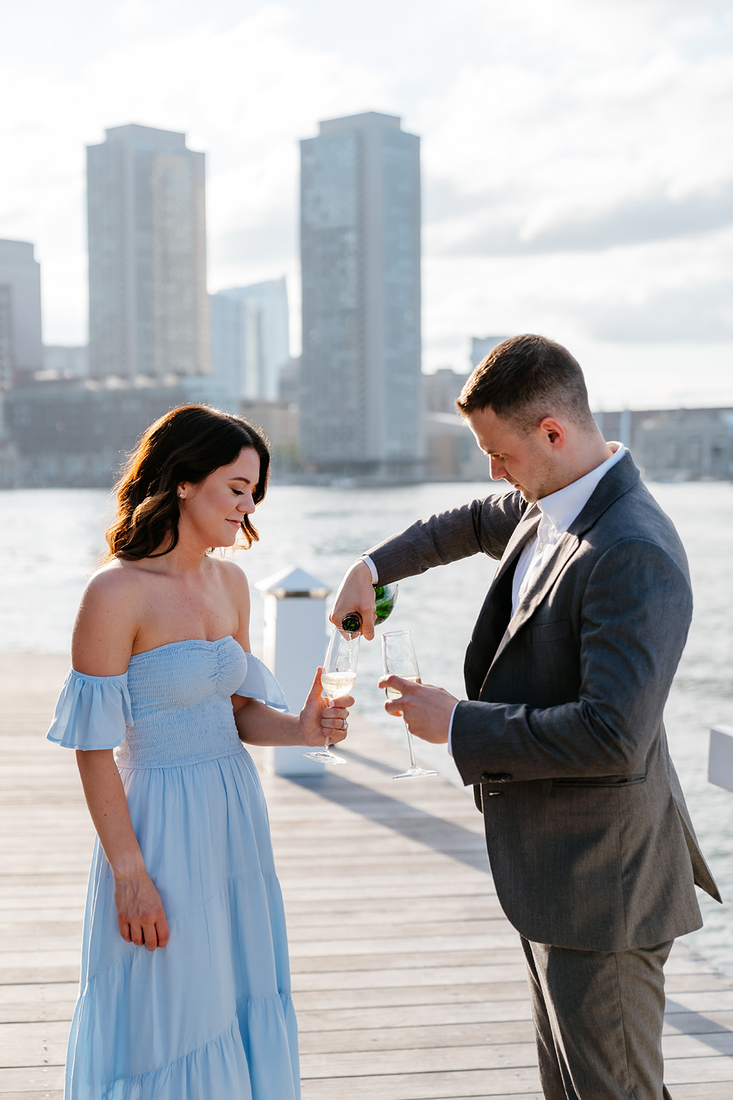 Man pouring champagne into woman's glass on a fan pier park with city skyline in the background as they take Boston engagement photos