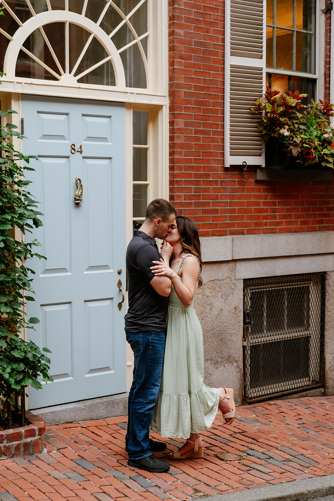 A couple embracing and kissing on a quaint city street with brick buildings and a blue door on the streets of Beacon Hill as they take Boston engagement photos