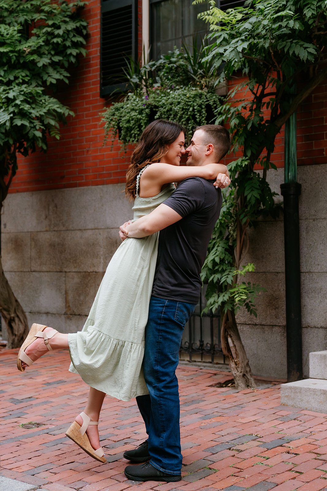 A couple embracing and smiling at each other on a brick sidewalk with greenery and a red brick building in the background on the streets of beacon hill 