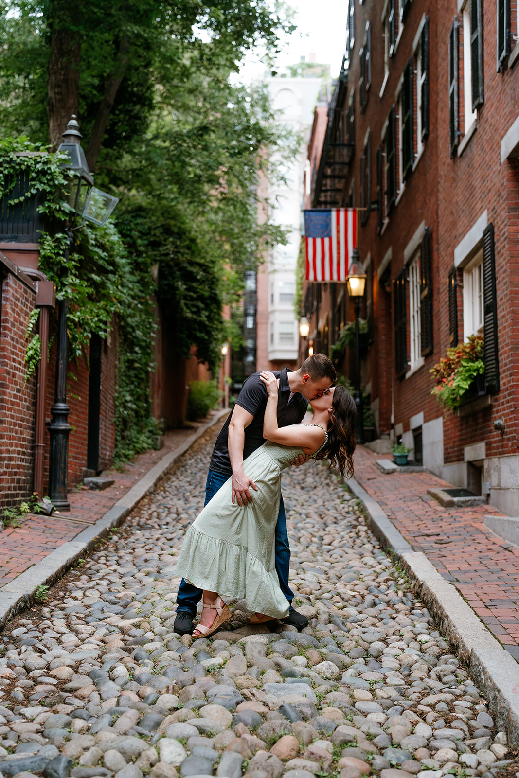A couple kissing in a cobblestone alley with brick buildings and an american flag in the background on the streets of beacon hill 