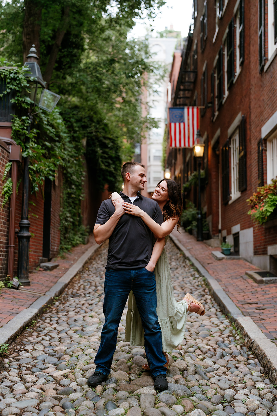 A couple embracing and smiling at each other on a brick sidewalk with greenery and a red brick building in the background on the streets of beacon hill