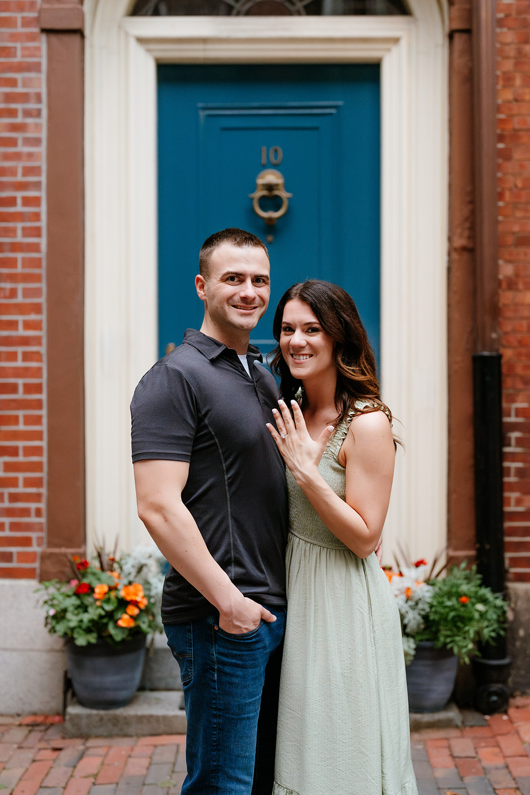 A couple embracing and smiling at each other on a brick sidewalk with greenery and a red brick building in the background on the streets of beacon hill 