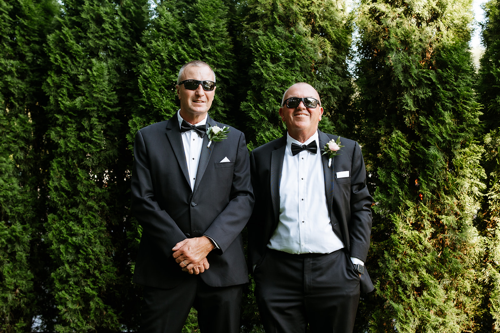 Two men in tuxedos with boutonnieres and sunglasses standing in front of tall green hedges.