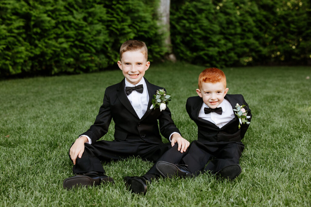 Two young boys in formal black tuxedos with boutonnieres sitting on a grass lawn with a hedge in the background.