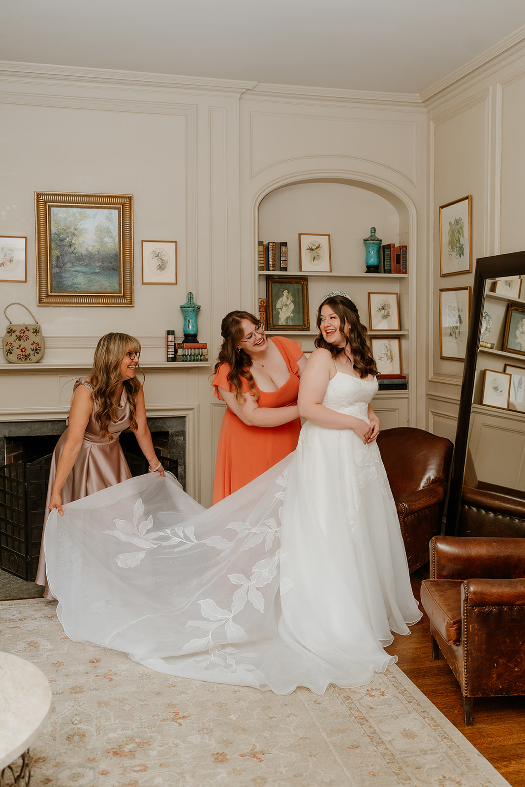 Three women in formal attire, one in a white bridal gown and two in dresses, smiling and adjusting the bride's dress in an elegant room at the estate at moraine farm