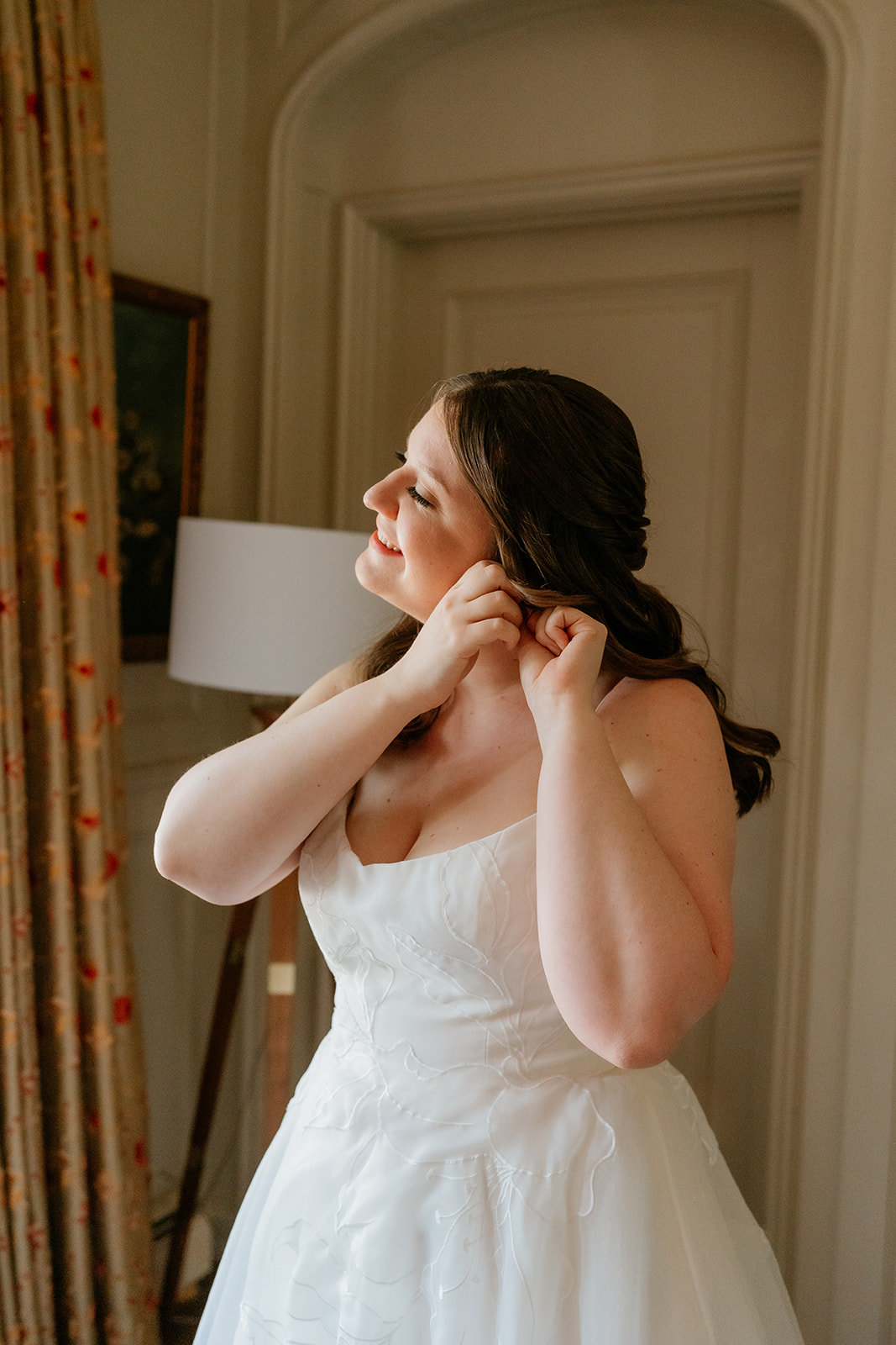A bride in a white dress fastens her earring while smiling and looking to the side in a well-lit room.