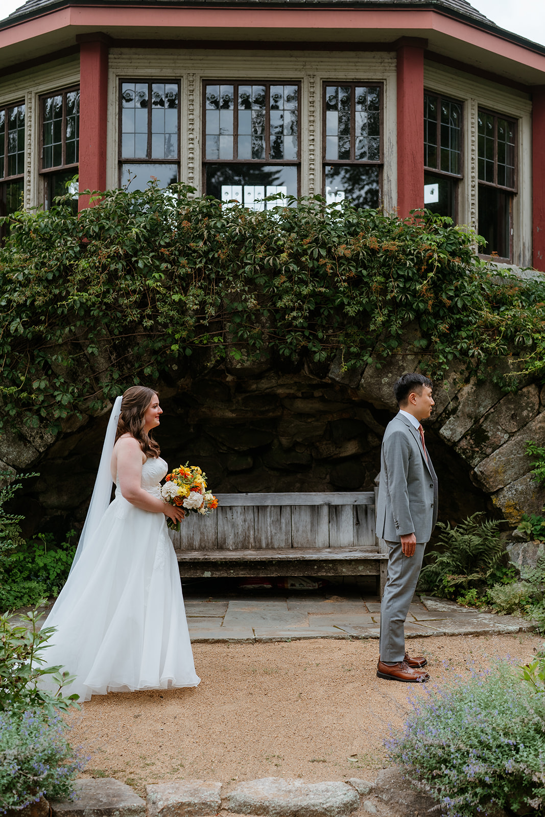 A bride and groom smiling at each other in a garden setting with a stone wall and wooden bench in the background at the estate at moraine farm