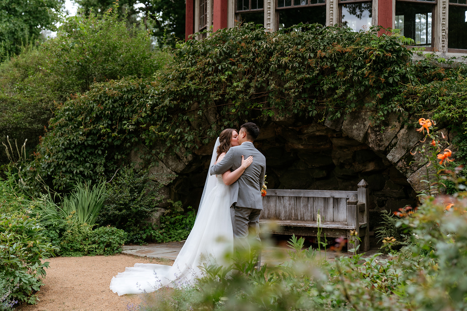 A bride and groom smiling at each other in a garden setting with a stone wall and wooden bench in the background at the estate at moraine farm
