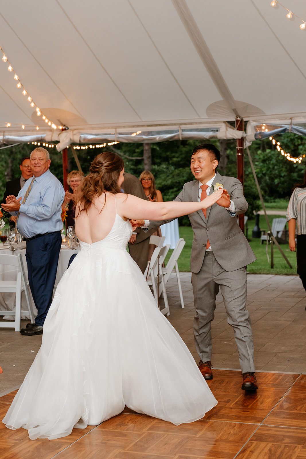 A bride and groom smiling and dancing under a tent with string lights, surrounded by guests.