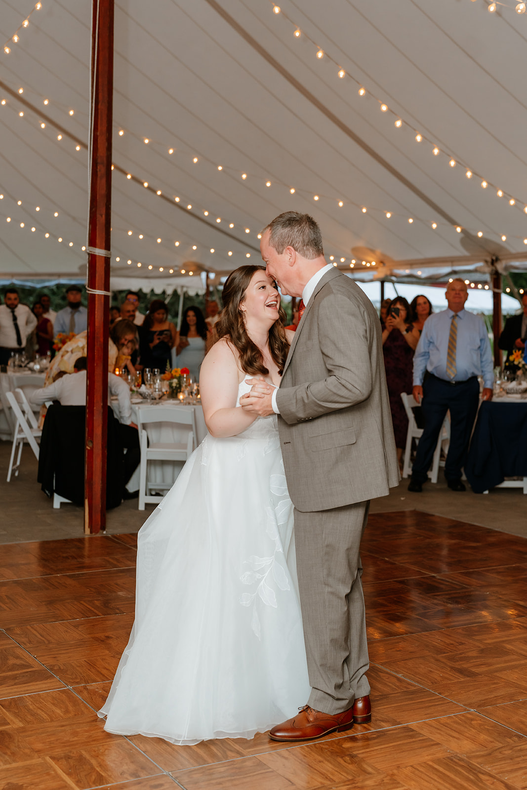 A bride and groom smiling and dancing under a tent with string lights, surrounded by guests.