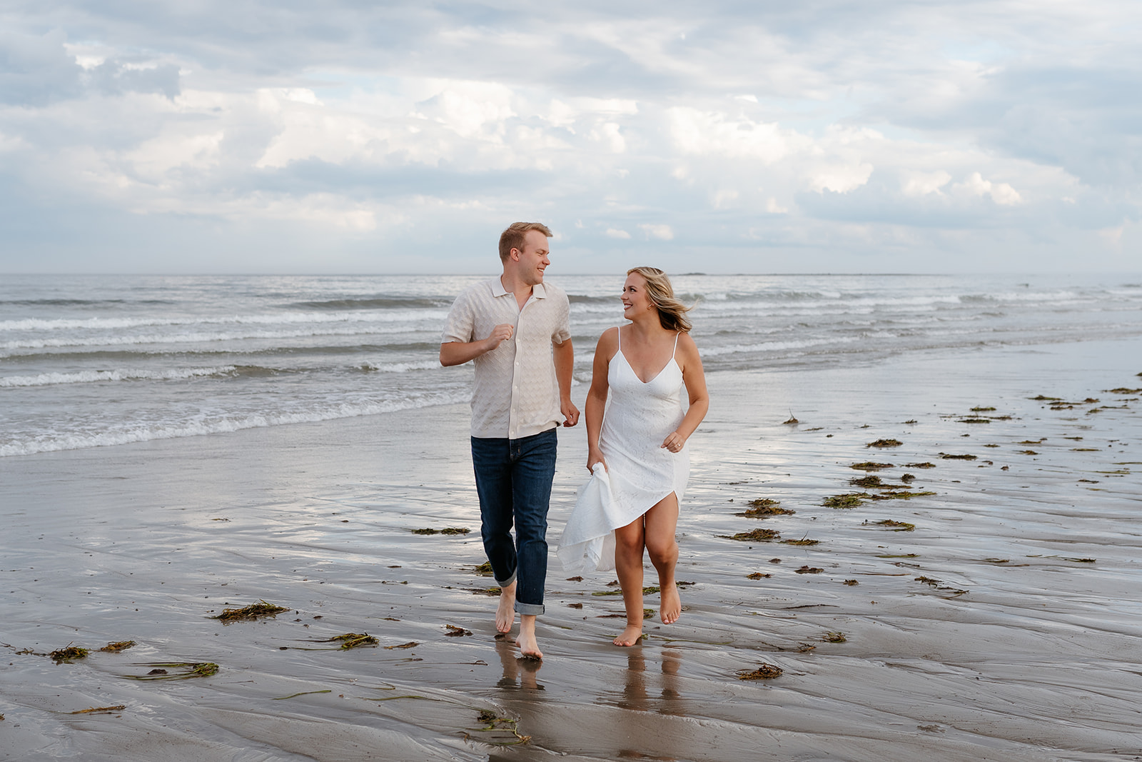 A couple holding hands and walking barefoot on a sandy beach, looking at each other, with cloudy skies in the background at their engagement session