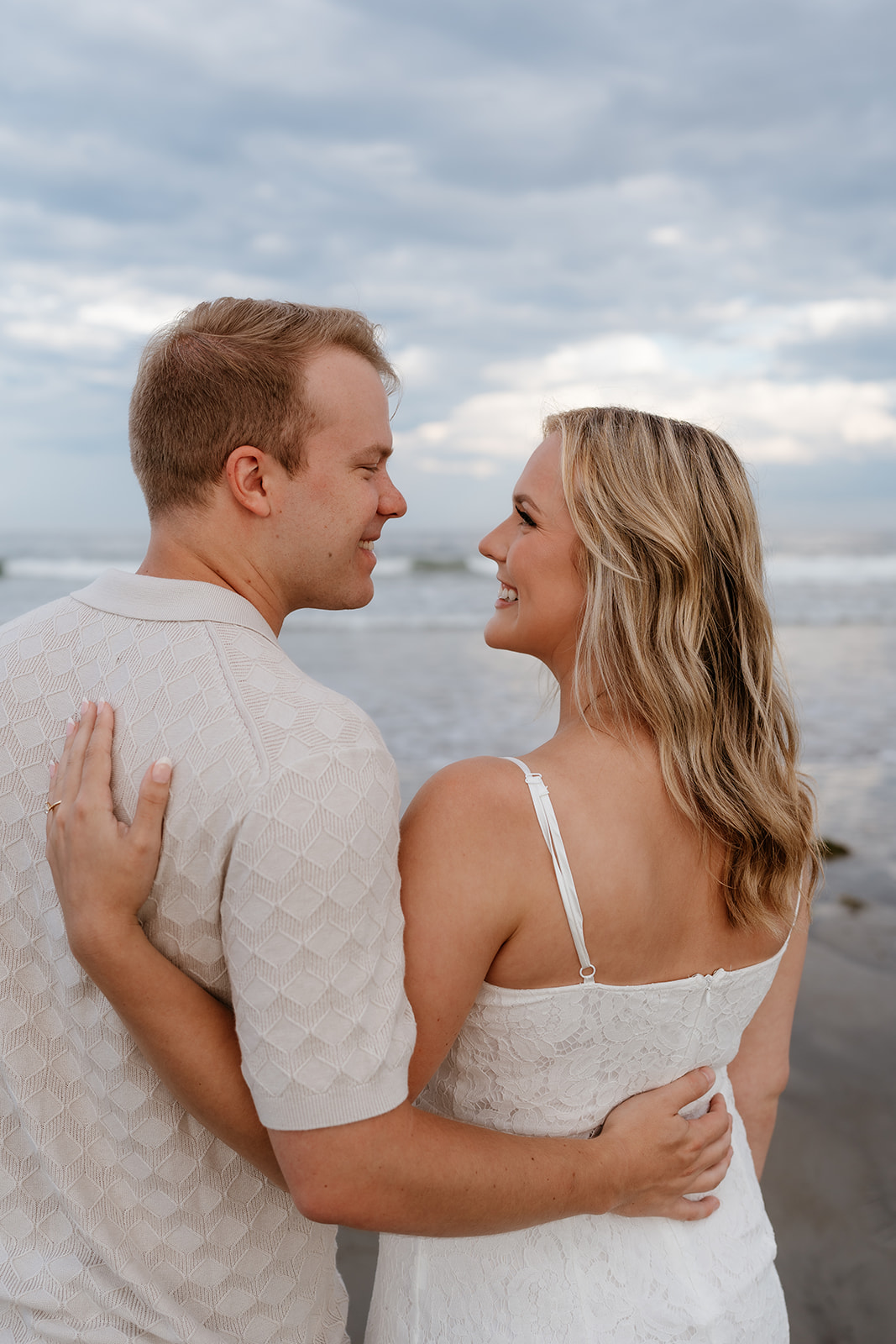 A couple holding hands and walking barefoot on a sandy beach, looking at each other, with cloudy skies in the background at their engagement session