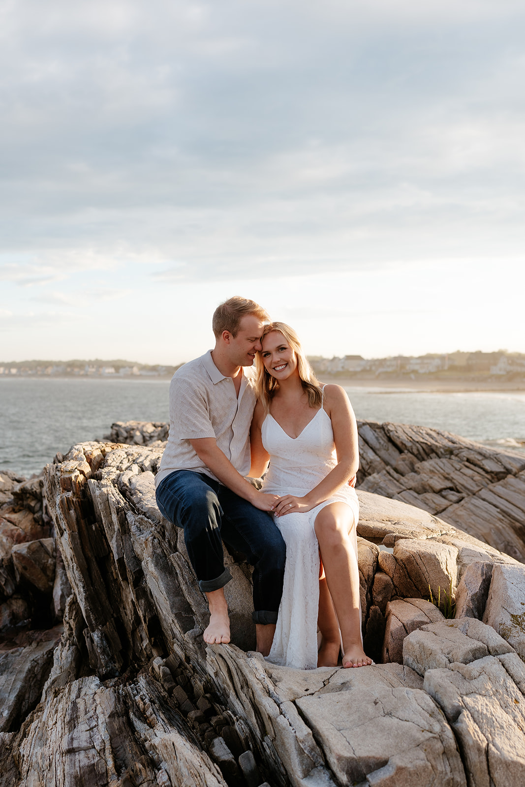 A couple sitting closely on rocky coastal terrain, smiling and embracing each other, with a serene water backdrop during sunset.