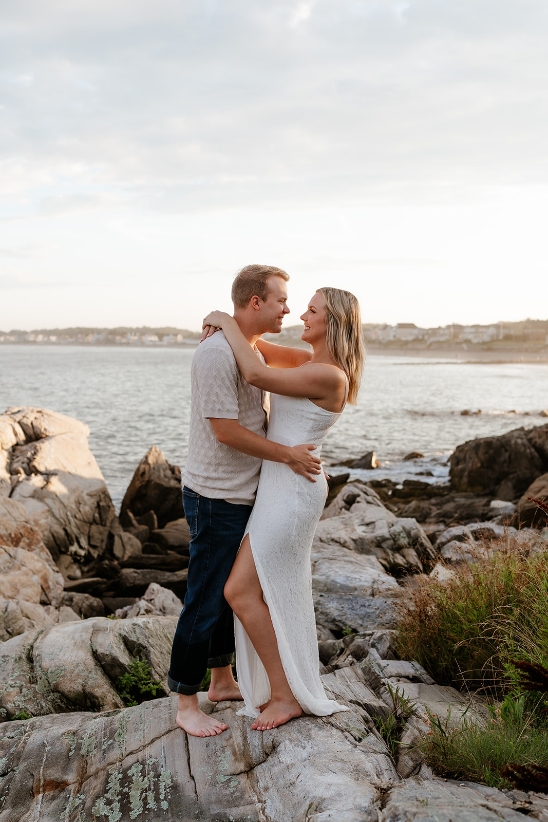A couple sitting closely on rocky coastal terrain, smiling and embracing each other, with a serene water backdrop during sunset.