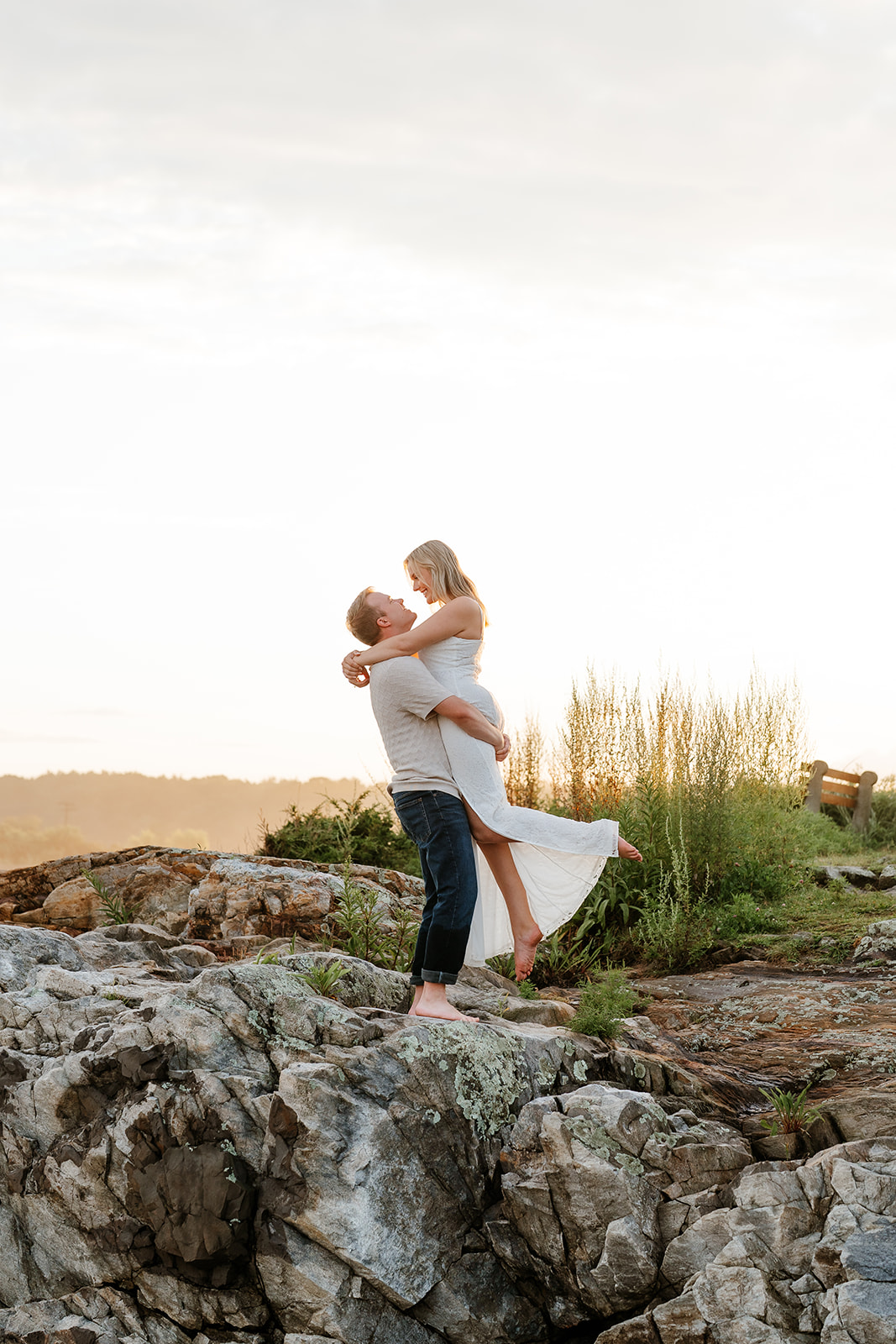 A couple playfully posing on a rocky coastline, with a woman in a white dress extending her arms like wings and a man in jeans behind her, both smiling at their beach engagement session