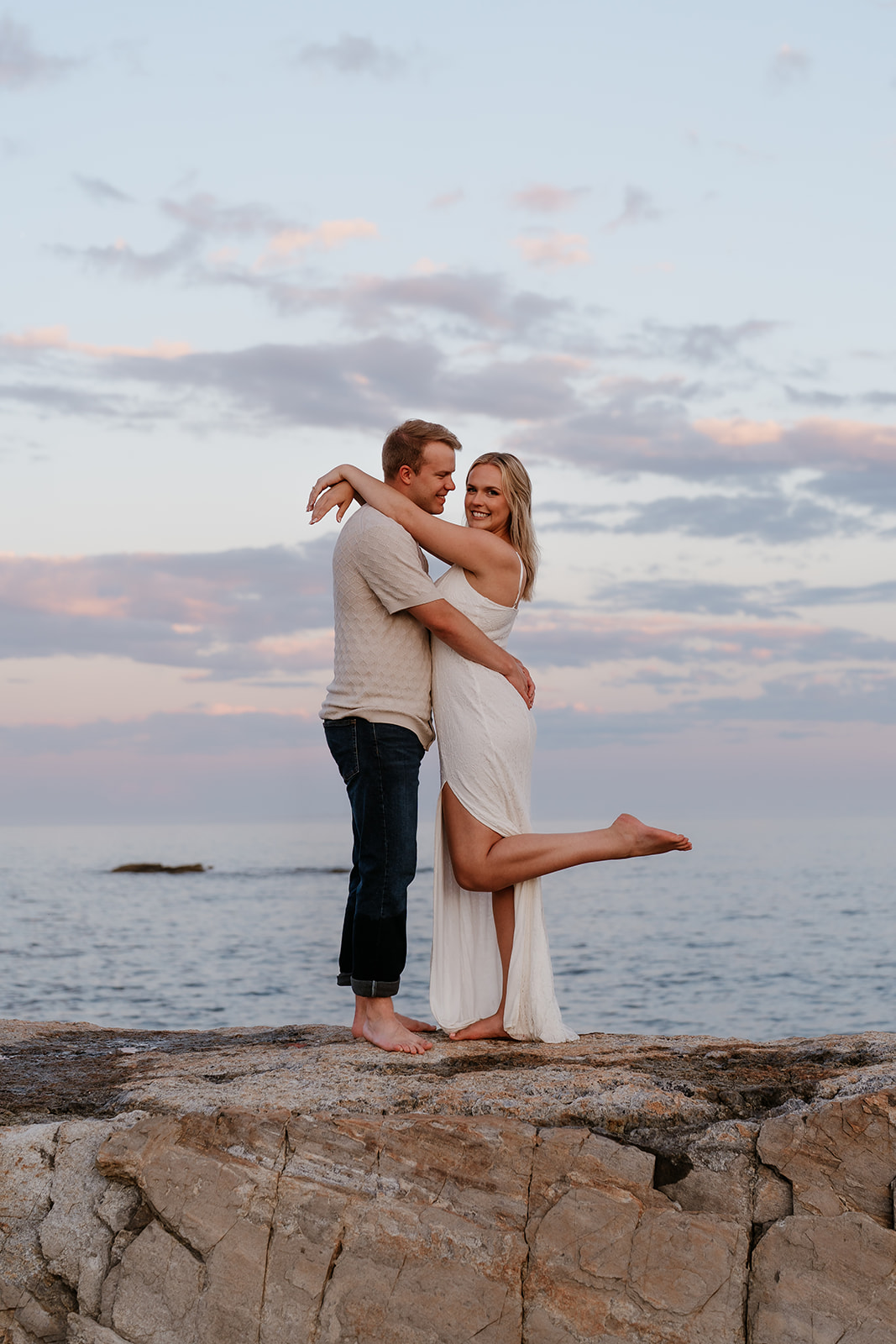 A couple playfully posing on a rocky coastline, with a woman in a white dress extending her arms like wings and a man in jeans behind her, both smiling at their beach engagement session