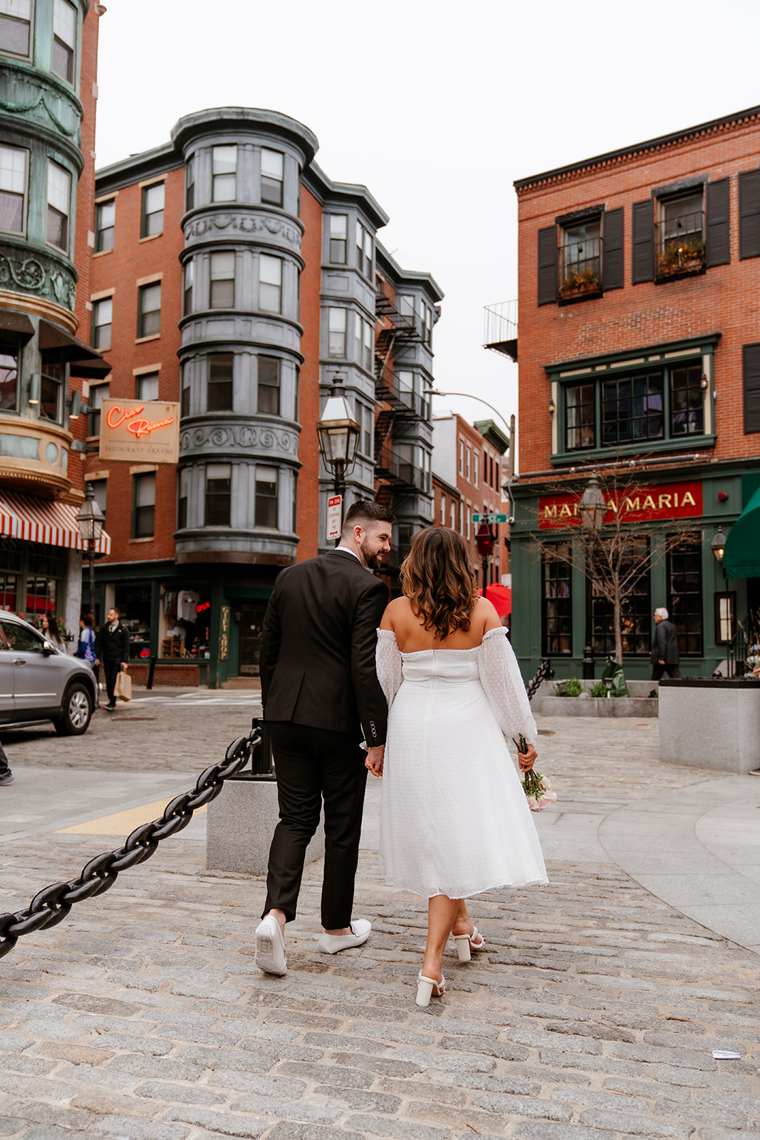 A couple embraces in a cobblestone street surrounded by red brick buildings in North End for their Engagement photos in Boston