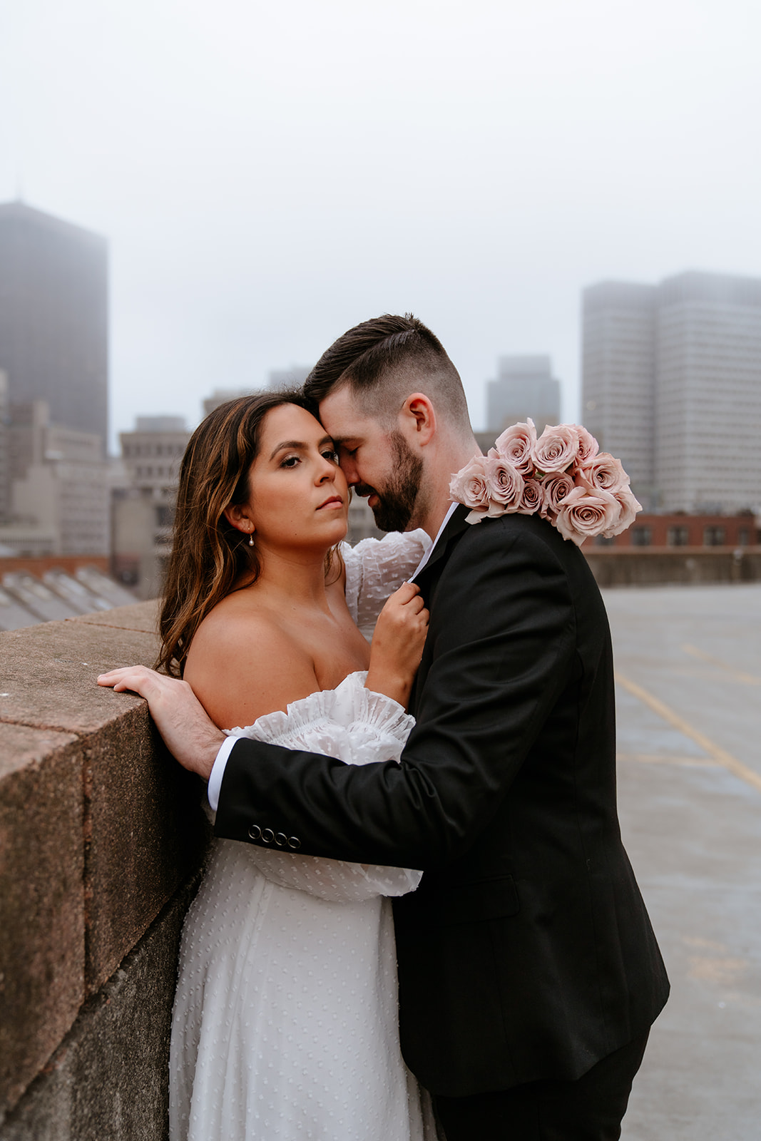 A couple stands on a rooftop parking garage in formal attire;Buildings and a foggy sky are visible in the background for their engagement photos
