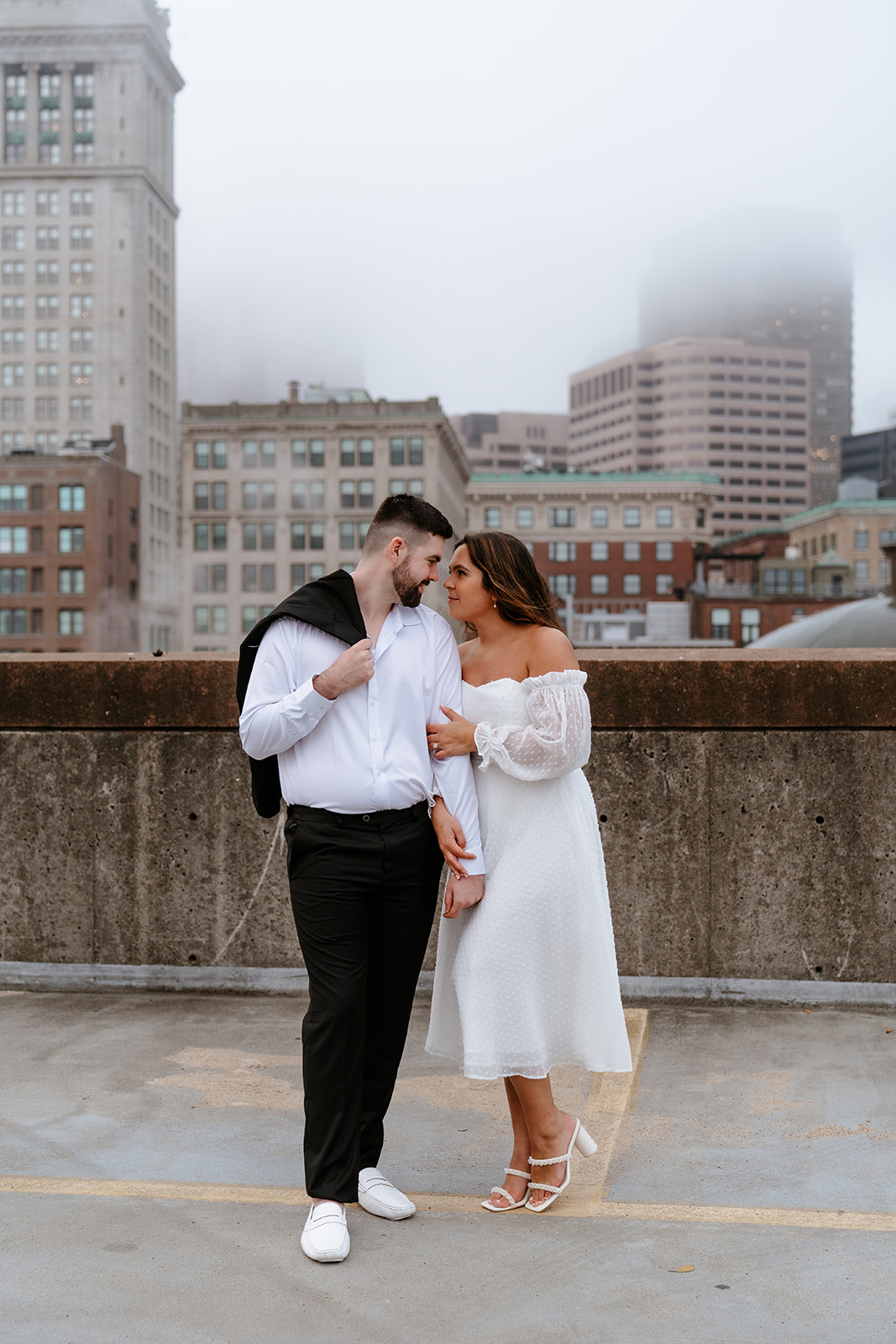 A couple stands on a rooftop parking garage in formal attire;Buildings and a foggy sky are visible in the background for their engagement photos
