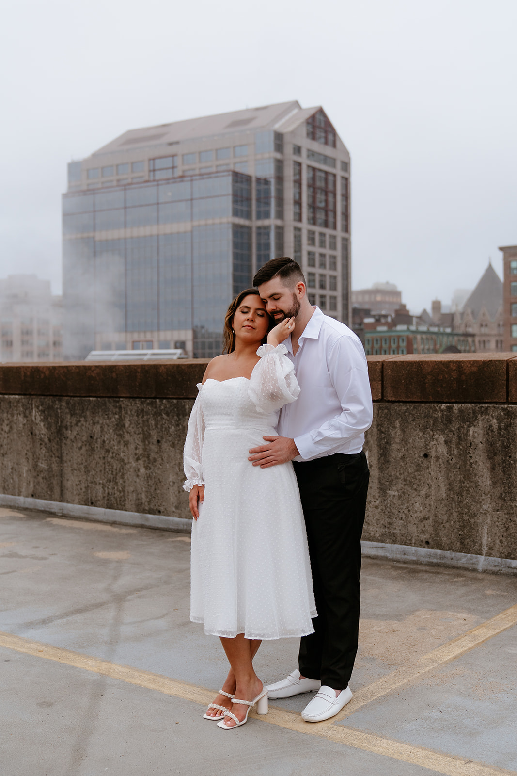 A couple stands on a rooftop parking garage in formal attire;Buildings and a foggy sky are visible in the background