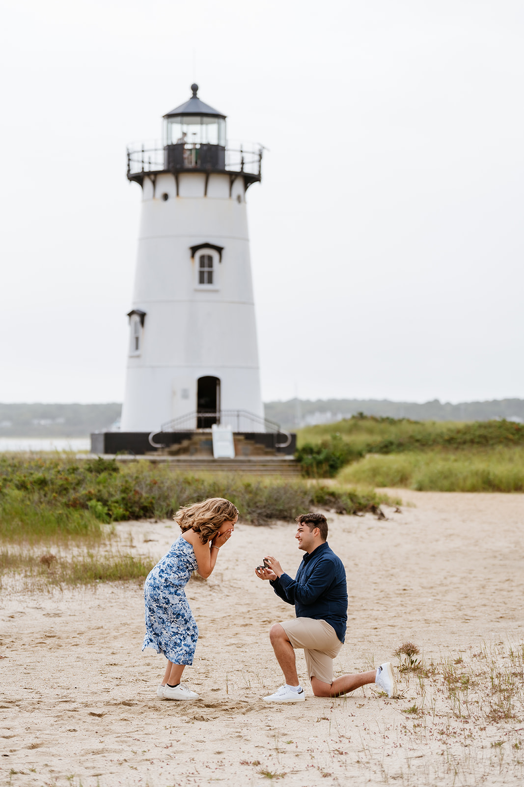 Man proposing to woman on one knee on sandy beach with Edgartown Harbor Lighthouse in the background.