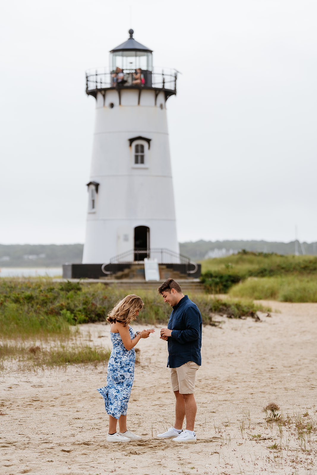 Man proposing to woman on one knee on sandy beach with Edgartown Harbor Lighthouse in the background.