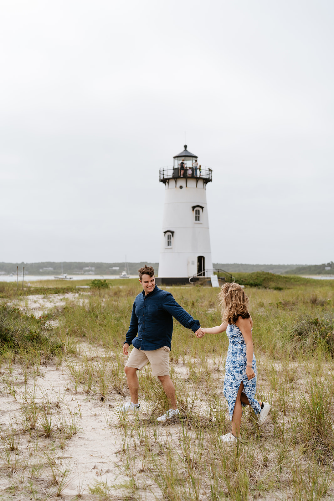 A couple kissing on a sandy beach with Edgartown Harbor Lighthouse  in the background. the woman wears a blue dress and the man a navy sweater and beige shorts.