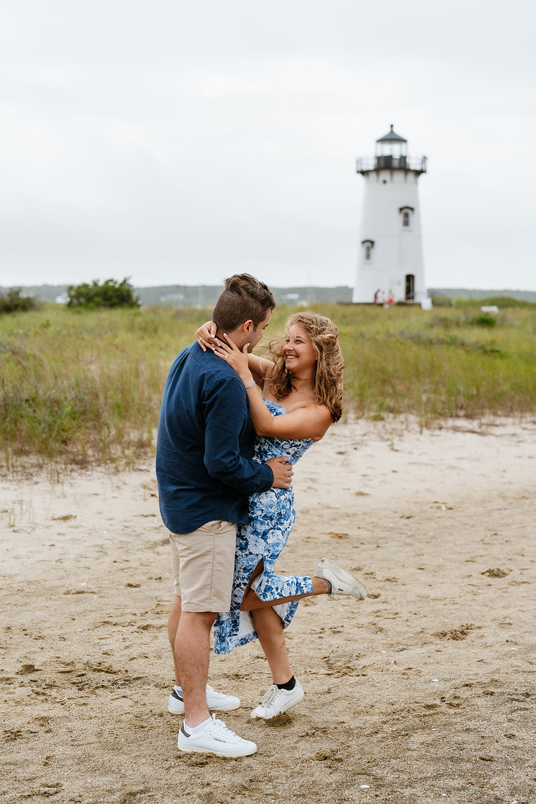 A couple kissing on a sandy beach with Edgartown Harbor Lighthouse  in the background. the woman wears a blue dress and the man a navy sweater and beige shorts.