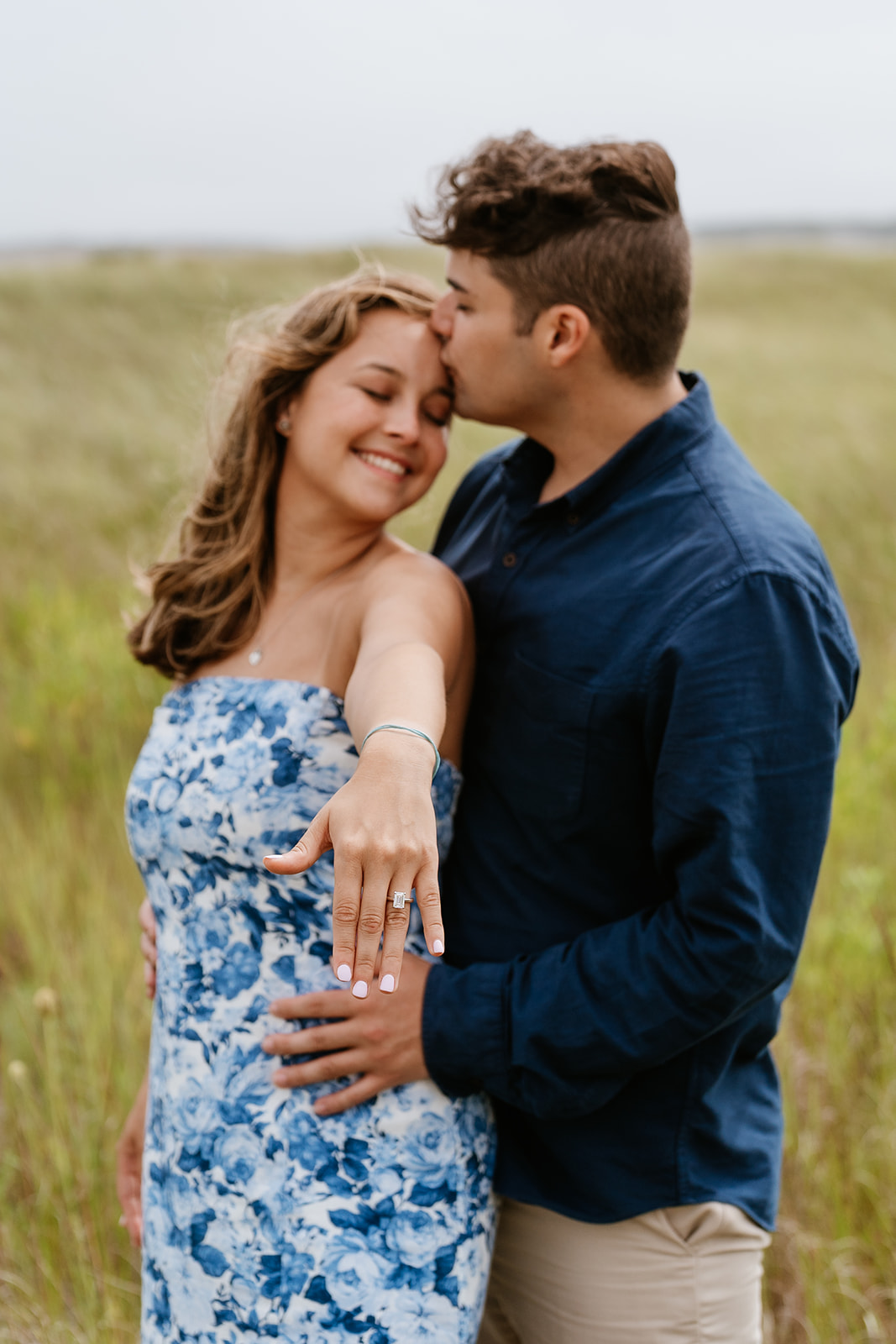 A happy couple embracing in a grassy field, with the woman showing off her engagement ring, both smiling towards the camera.