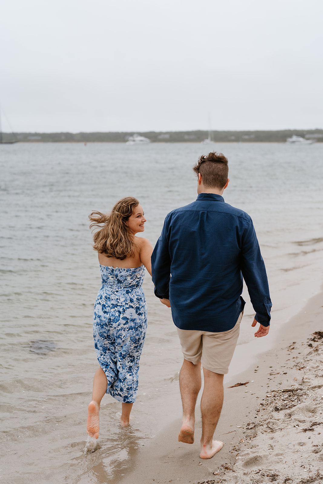 A couple holding hands and running barefoot along a sandy beach in Martha's VIneyard with boats in the background.