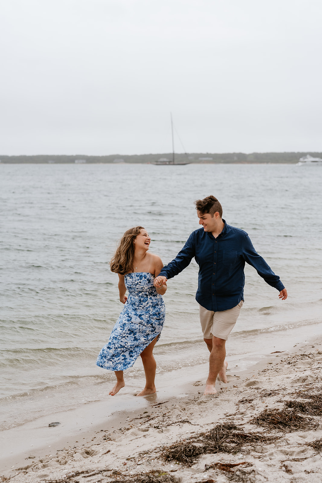 A couple holding hands and running barefoot along a sandy beach in Martha's Vineyard with boats in the background.
