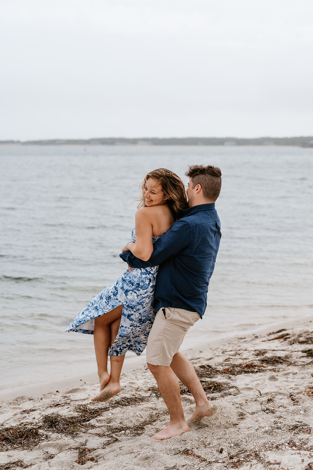 A couple holding hands and running barefoot along a sandy beach in Martha's Vineyard with boats in the background.