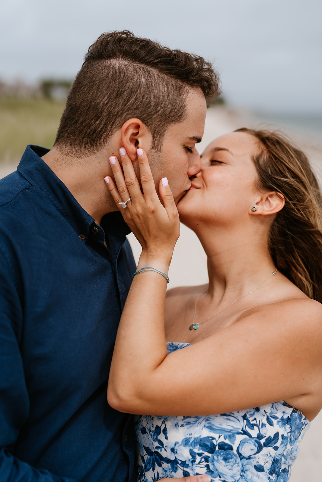 A couple kissing along a sandy beach in Martha's Vineyard with boats in the background.