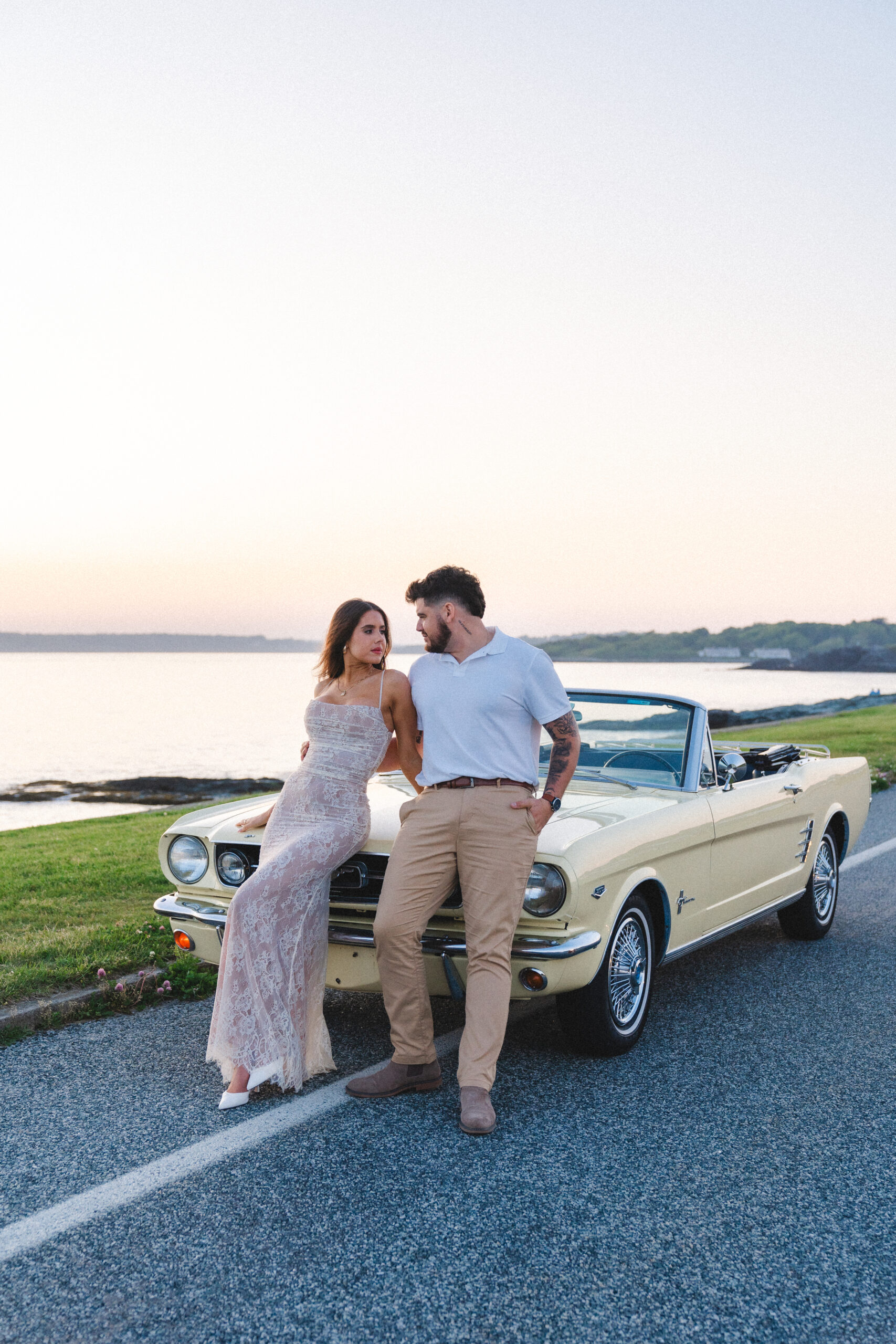 Newly engaged couple smiling at eachother standing in front of a classic car