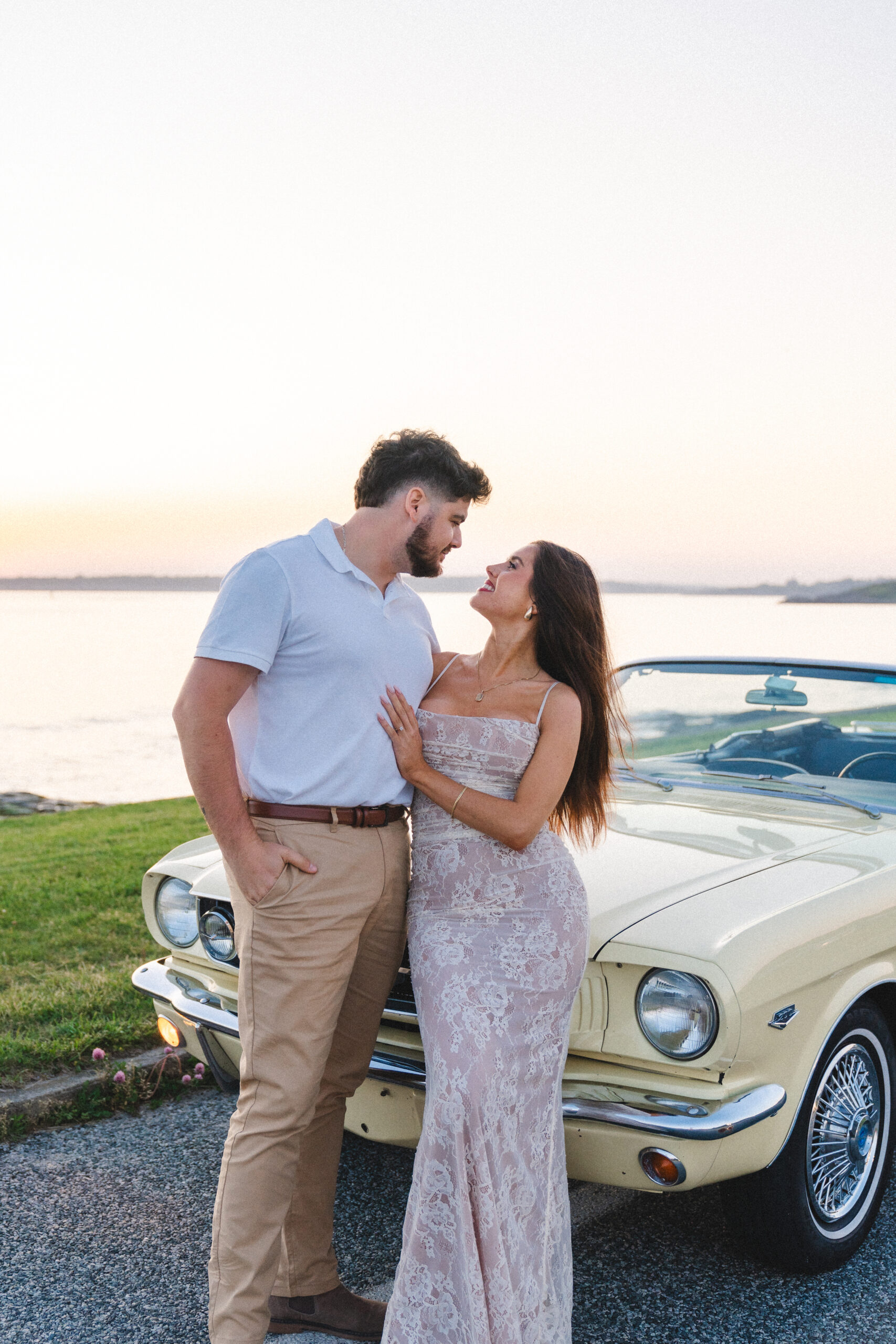 Newly engaged couple smiling at eachother standing in front of a classic car