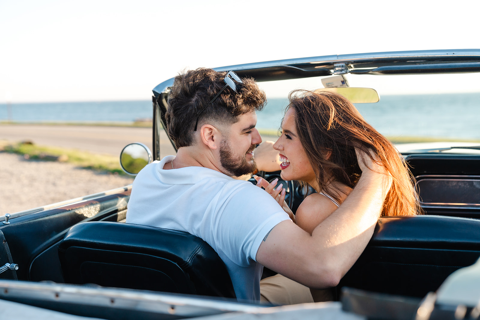 Newly engaged couple smiling and laughing at one another sitting in a classic car