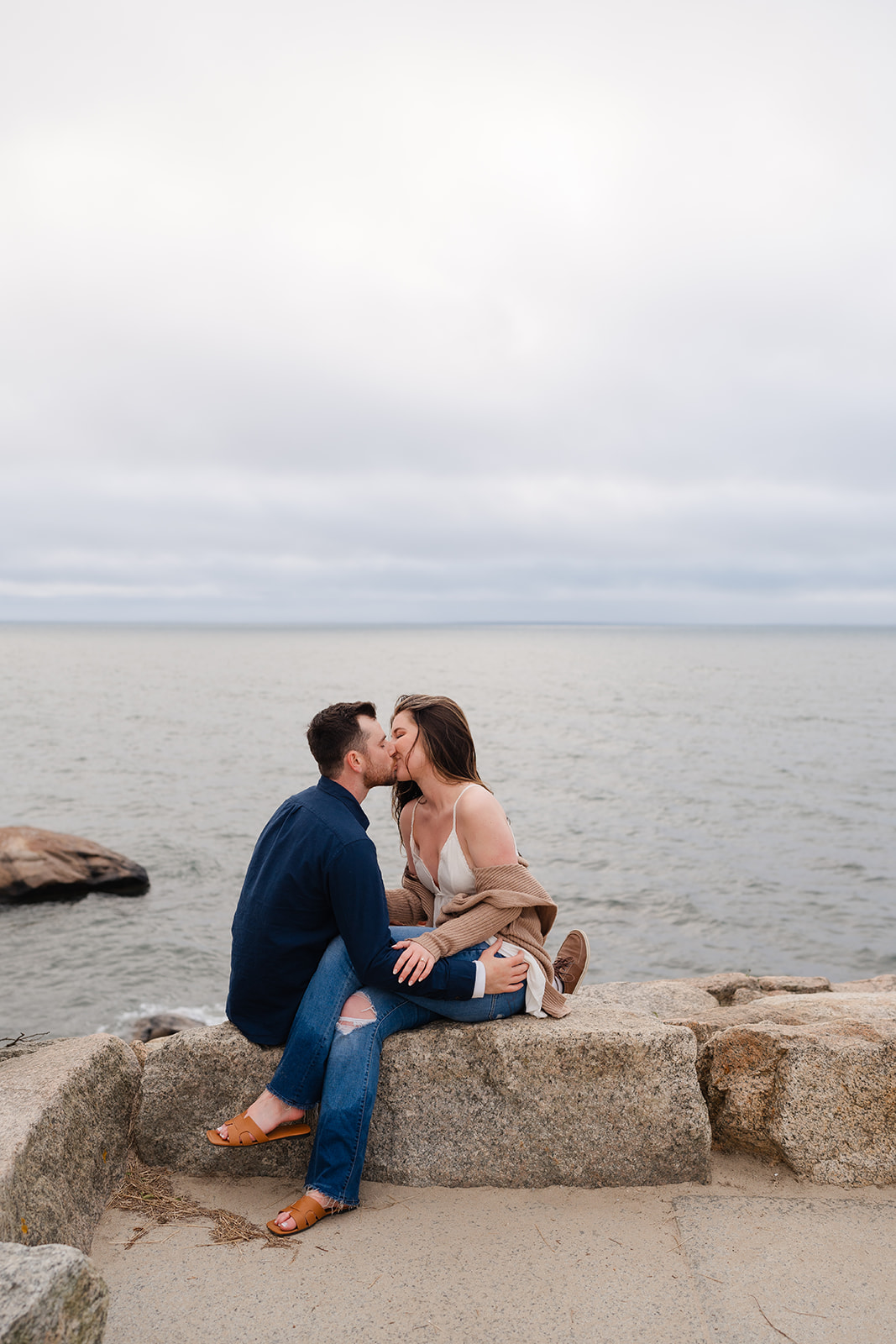 Engaged couple sitting on a brick wall at The Knob at Cape code kissing