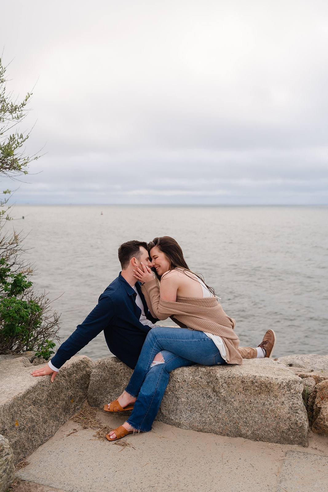 Engaged couple sitting on a brick wall laughing smiling at one another during their Cape Cod engagement 