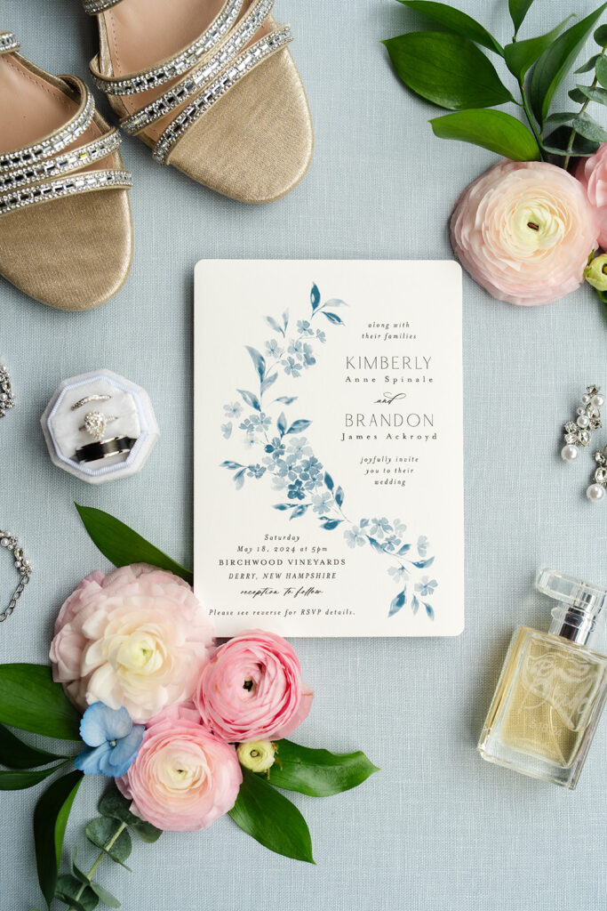 Flat lay of bride's items: shoes, perfume, earrings and rings with pink flowers and the wedding invitation