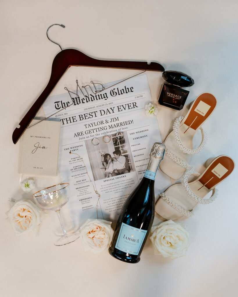 Wedding invitation as a newspaper for their flat lay with white roses, champagne bottle and glass, perfume and bride's pearl heels