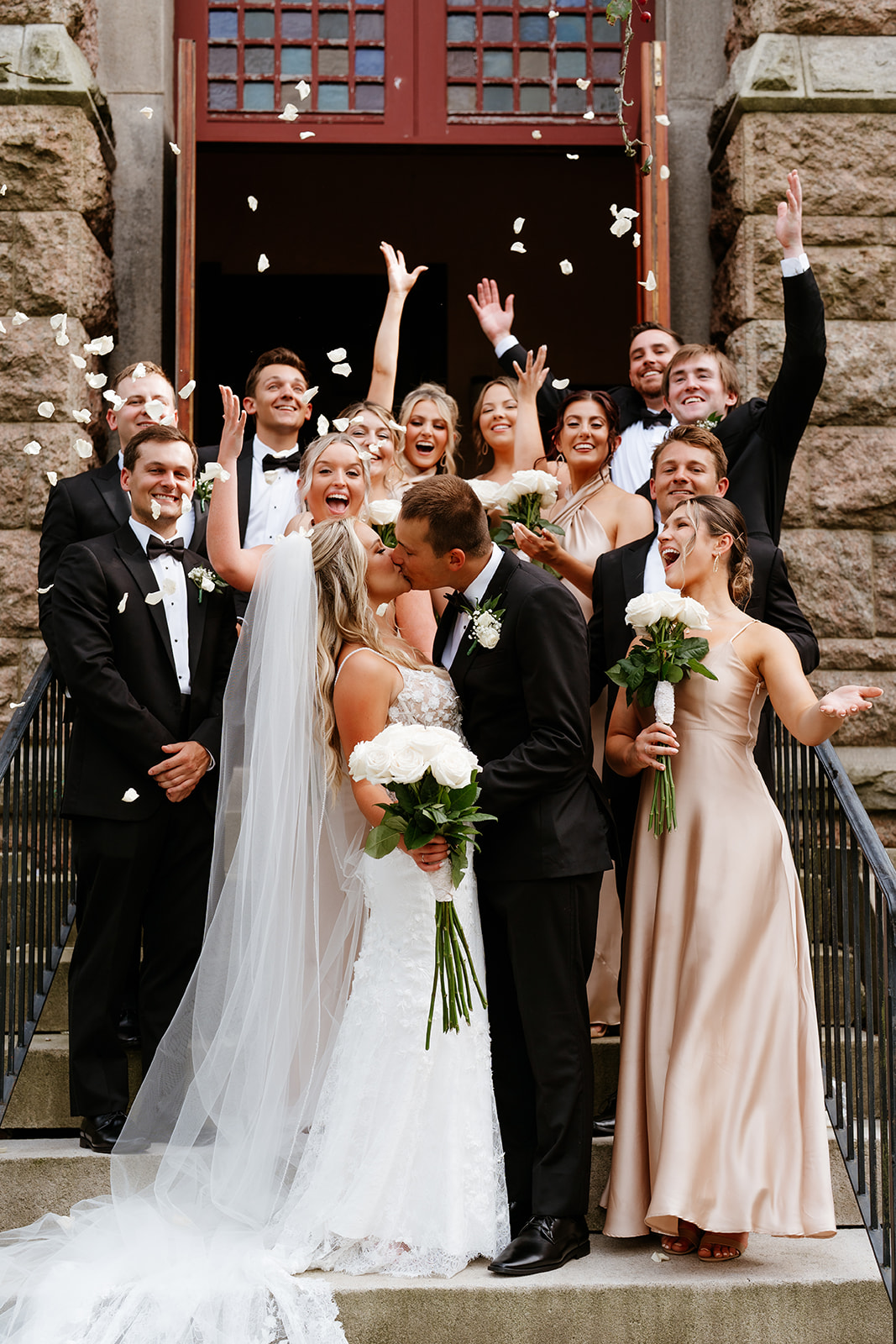 bride and groom kisses in front of the church with bridesmaids and groomsmen throwing rose petals