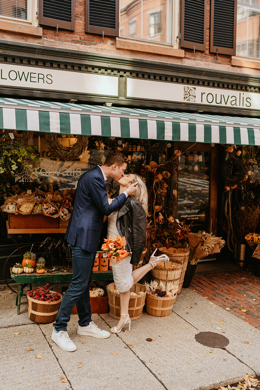 engaged couple kissing while holding an autumn bouquet in front of florist shop