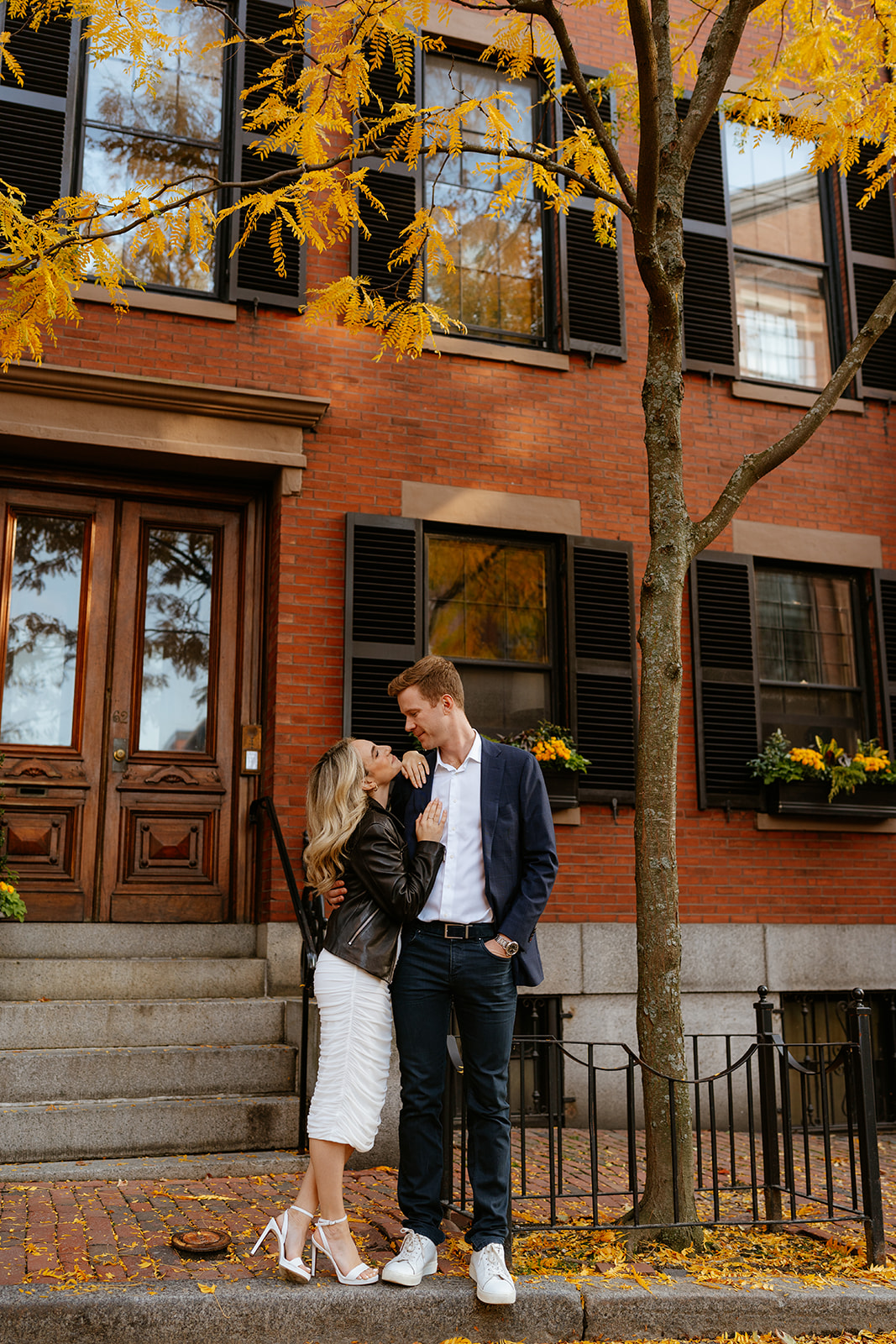 engaged couple looking at each other with brick apartment background with fall leaves 