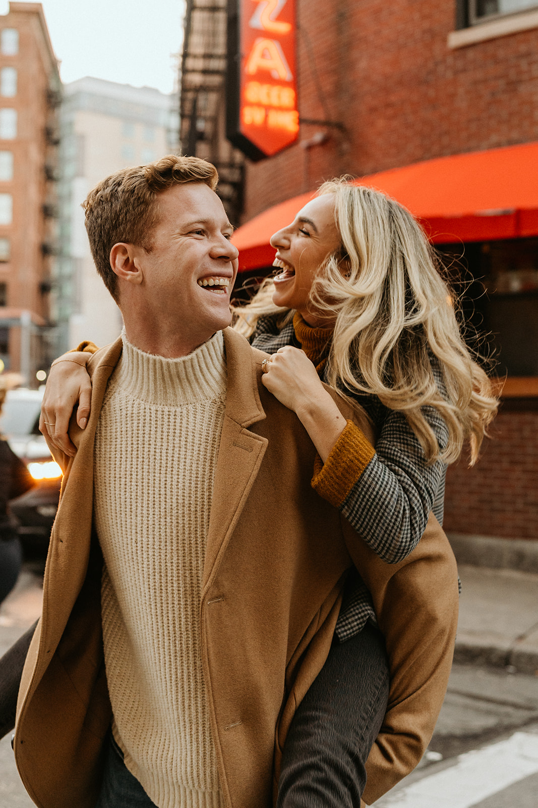 engaged couple smiling and laughing while piggy backing wearing winter outfits 