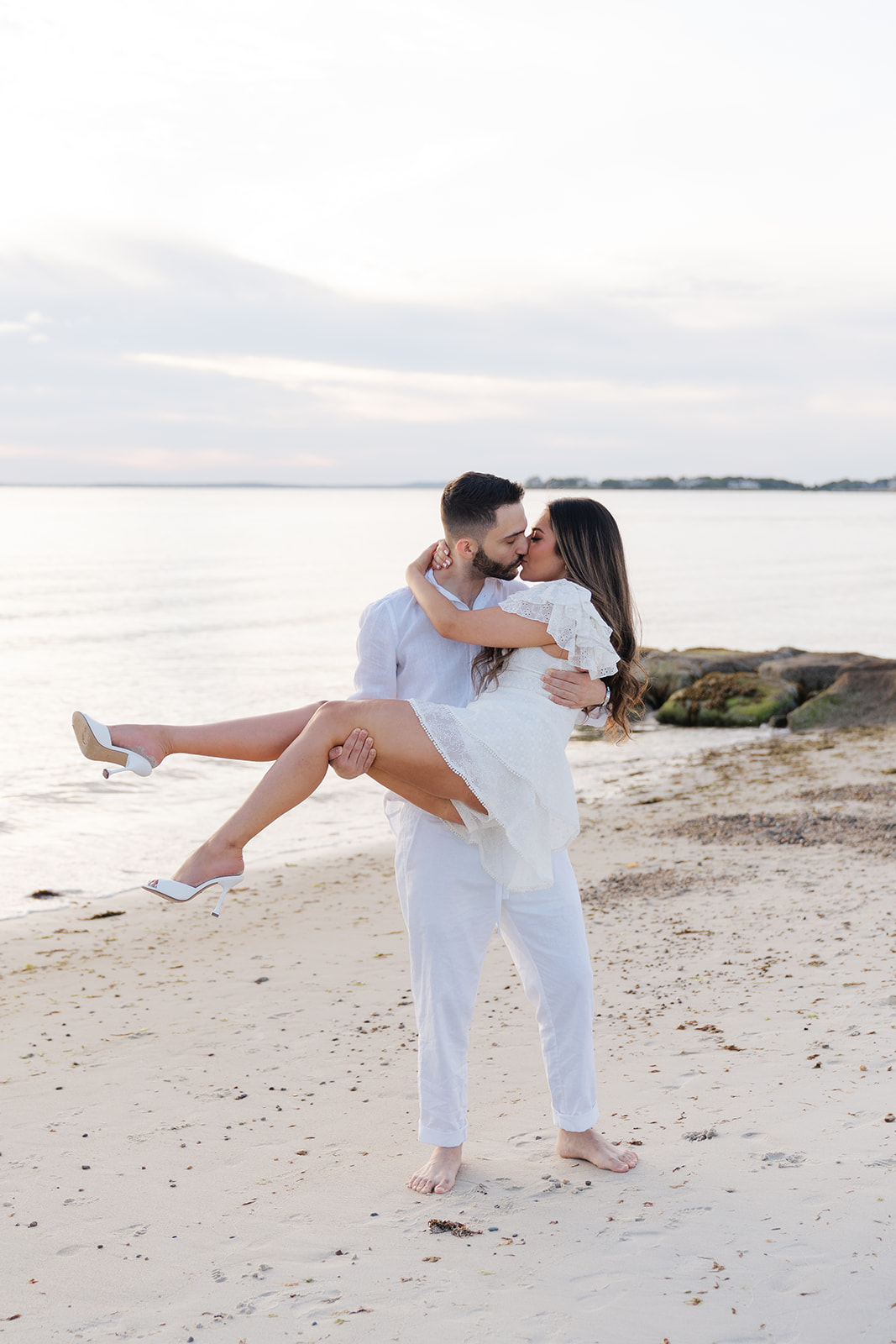 The couple sharing a playful and intimate moment with one partner lifting the other in the air on the beach at sunset, a stunning example of Cape Cod engagement photos.