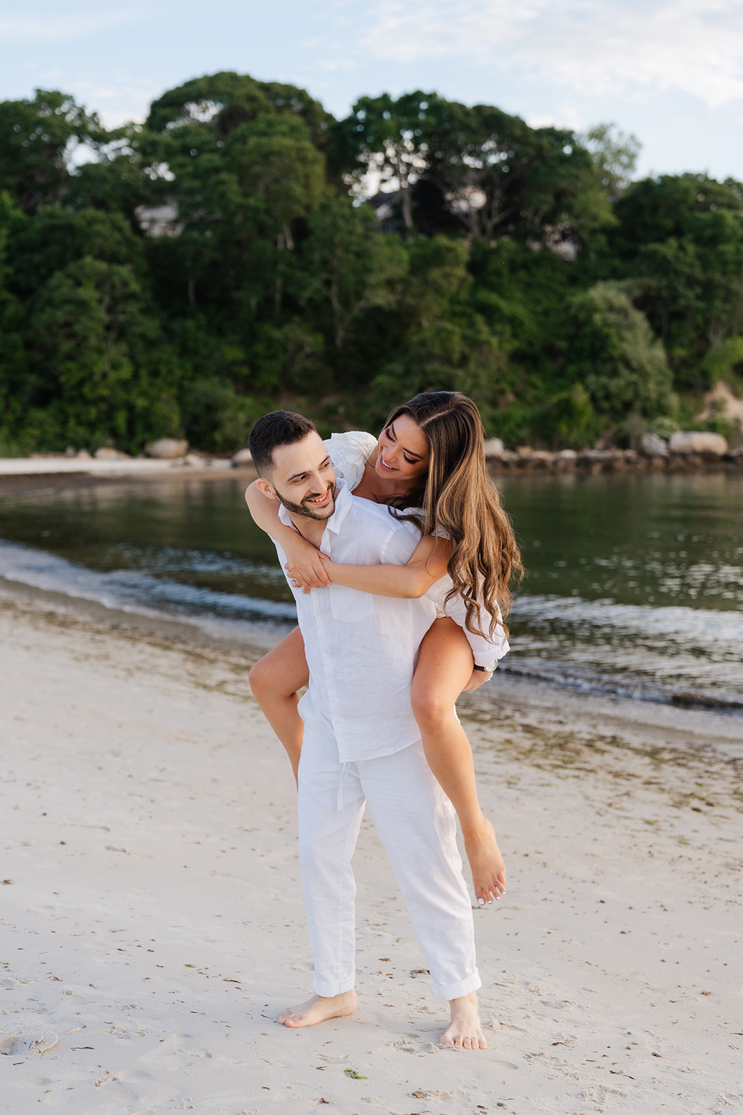 A loving and joyful moment where the couple shares a piggyback ride on the beach, with smiles and laughter as the ocean gently waves in the background, an ideal setting for Cape Cod engagement photos.