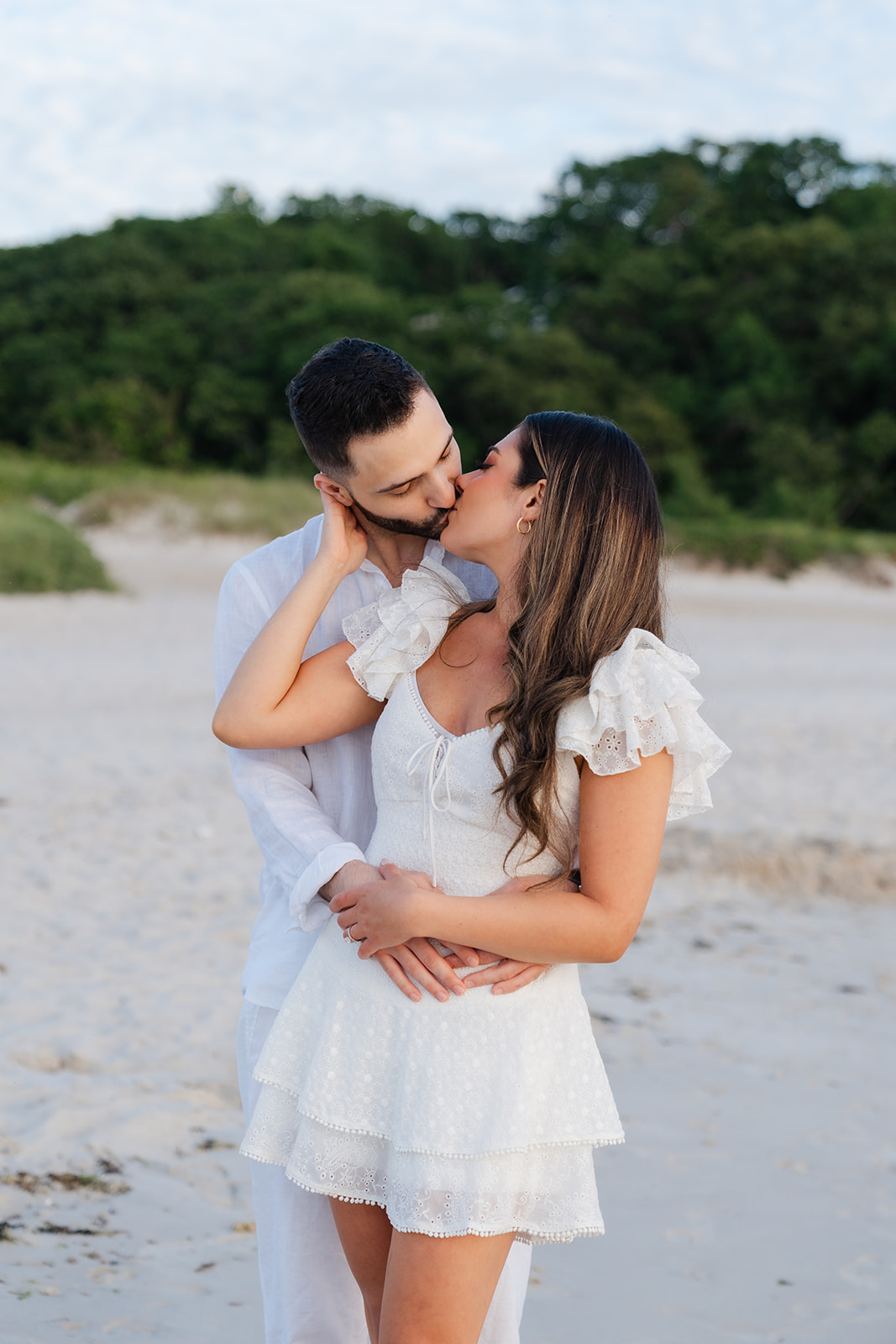 A close-up of the couple sharing an intimate kiss on the beach at sunset, with the soft lighting and tranquil water setting the mood for a memorable Cape Cod engagement photo.